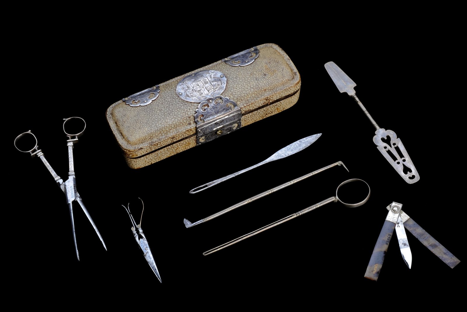 An ornate gold pocket case from the 17th century, on a black background, surrounded by the instruments it held: scissors, a scalpel, fine probes, a hollow spatula and a knife with hinged blades.