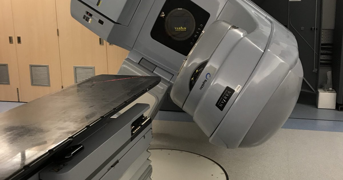 Acelerador linear de radioterapia cinza inclinado sobre a mesa de tratamento numa sala de hospital