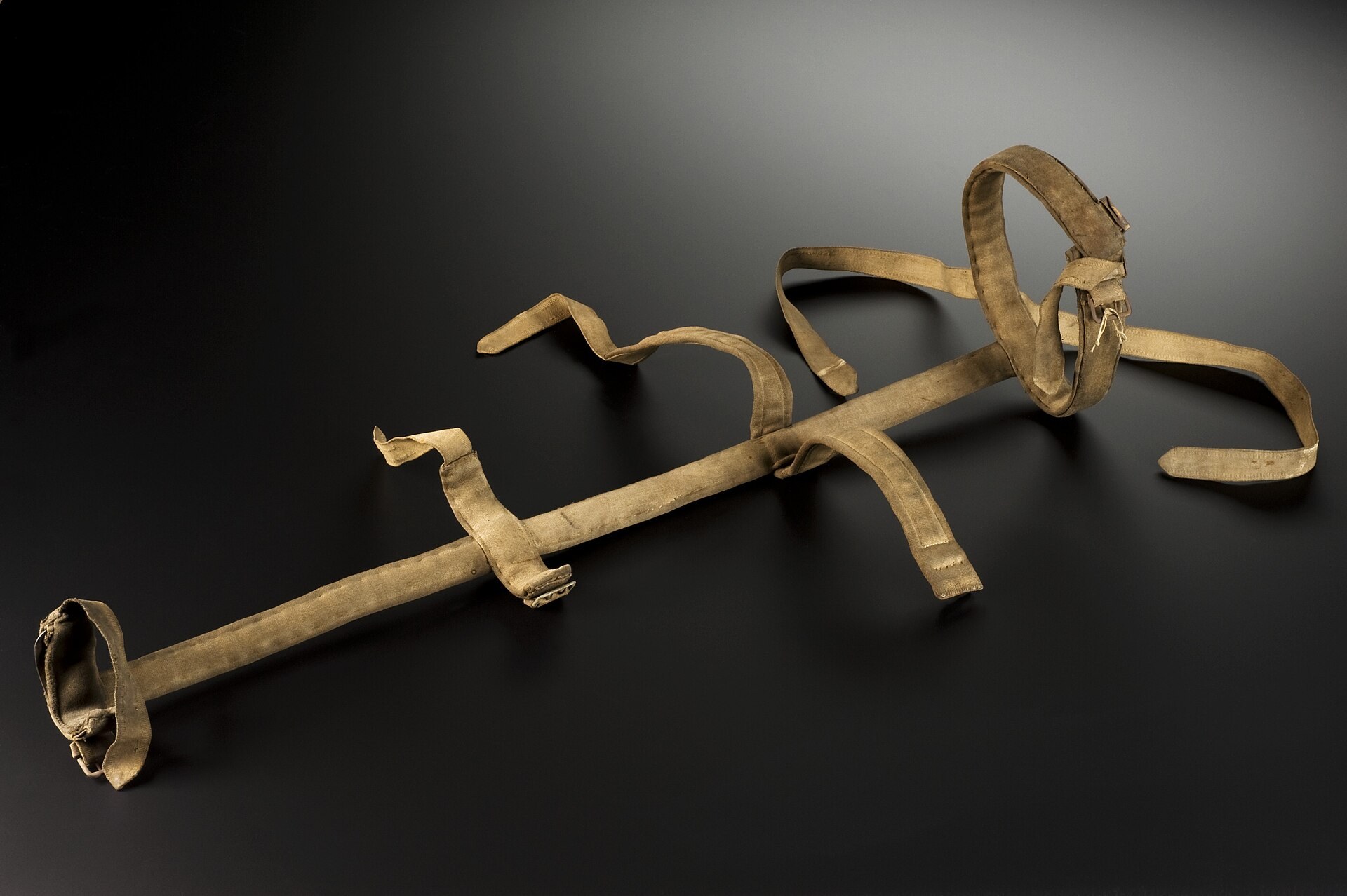 Attelle de jambe ancienne de type Thomas, une longue tige de métal garnie de tissu beige, avec un anneau rembourré à une extrémité et plusieurs lanières de fixation détachées sur toute la longueur, sur fond sombre.