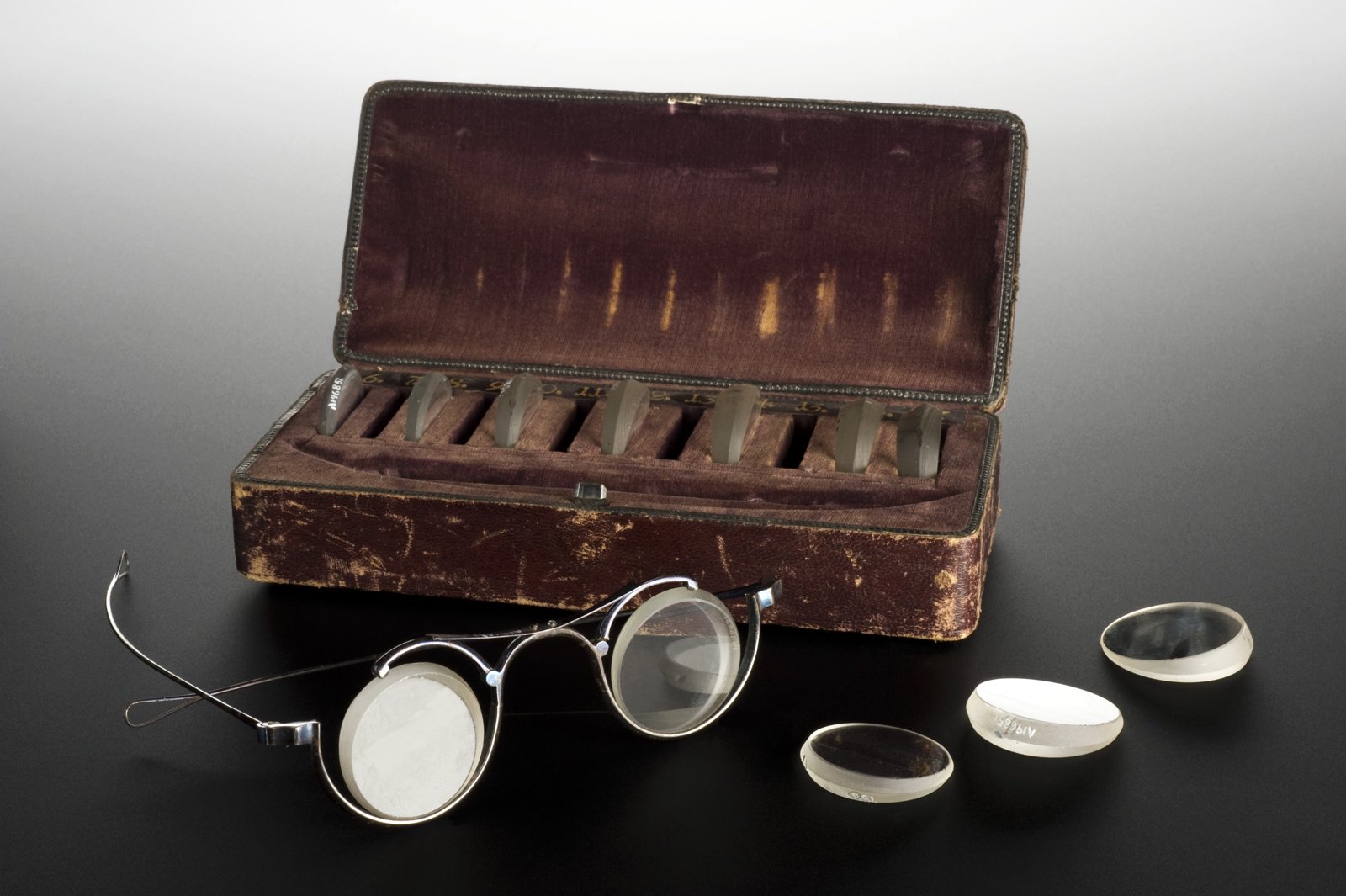 Old case lined with wine-colored velvet, open, with a row of trial lenses fitted in place, a metal trial frame with two lenses in front and three loose lenses on a dark background.