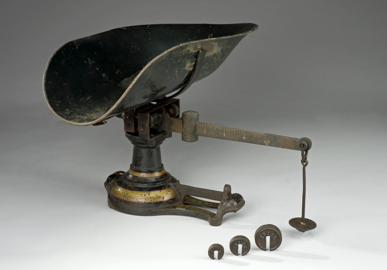 Old baby weighing scale, made of iron painted black with gold bands, with a deep bowl-shaped pan, a graduated arm with a plate hanging from the end and three small weights on the floor, on a light gray background.