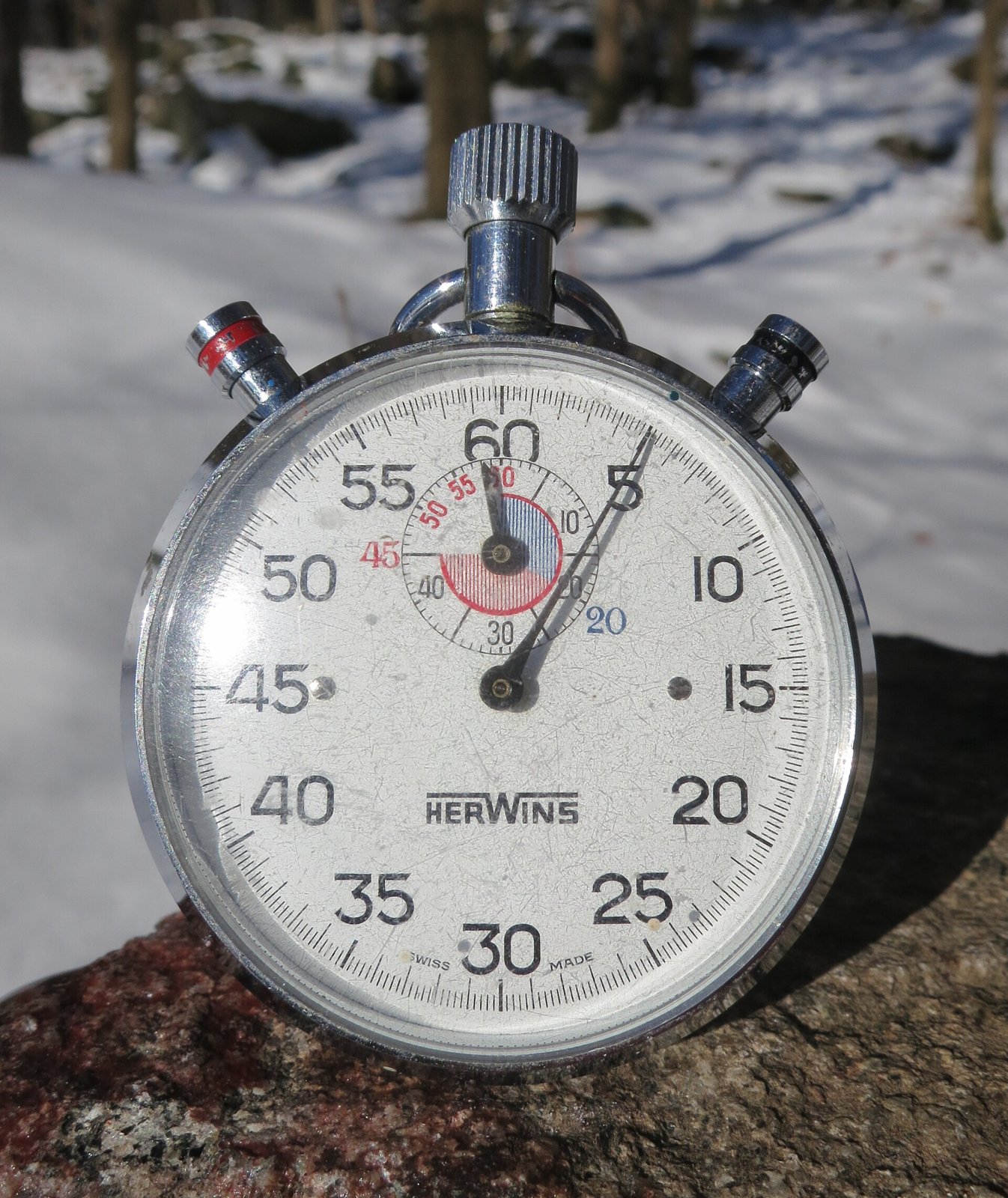 Old mechanical pocket stopwatch, with a white dial numbered every five seconds, black hand and metal crown, resting on a stone, with snow and trees in the background.