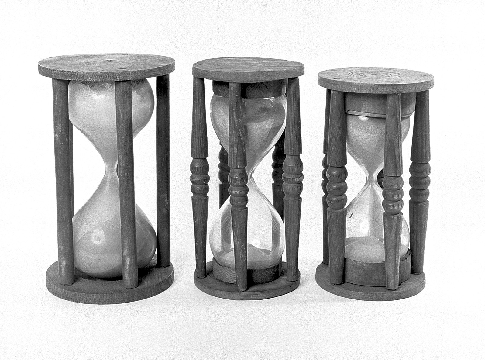 Three old wooden and glass hourglasses, side by side, photographed in black and white on a light background, with the sand gathered in the lower bulb.