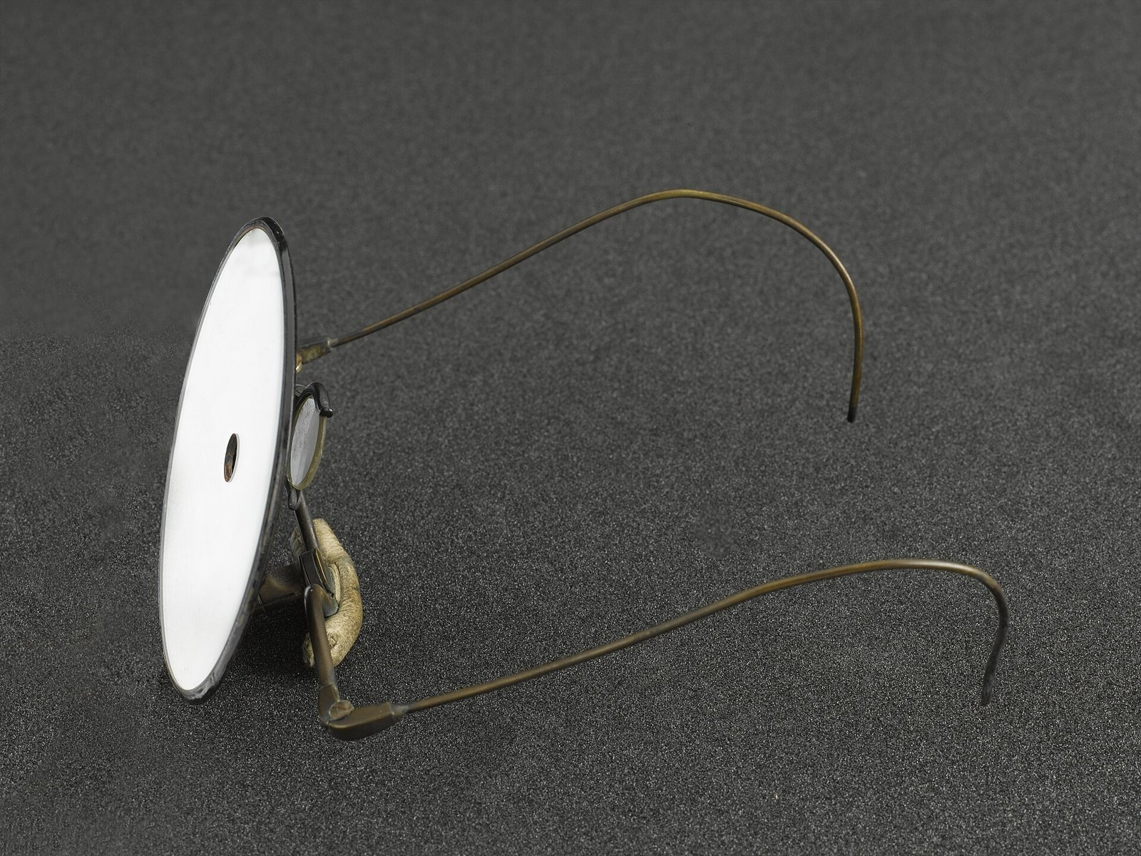 Old reflecting laryngoscope: a round white mirror with a hole in the center, attached to a pair of glasses frames with thin metal arms, photographed on a dark gray background.