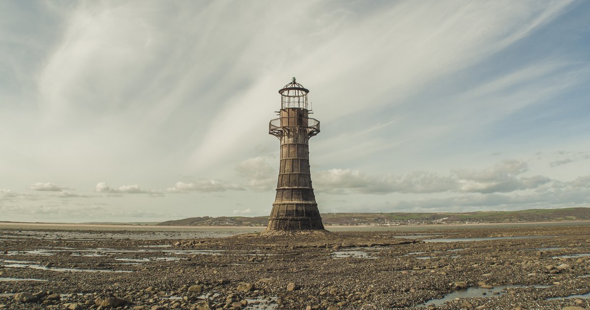Farol de Whiteford, uma torre de ferro escura, isolado sobre a areia e as pedras da maré baixa sob um céu de nuvens finas.