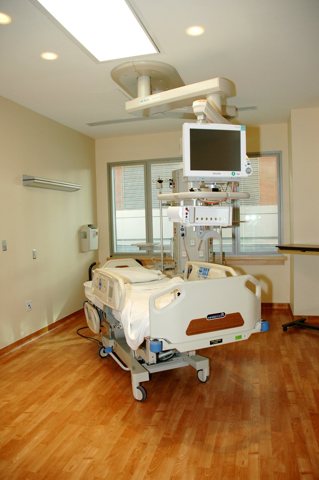Leeres Krankenzimmer mit hellem Holzboden, einem Bett mit weißem Laken und einem ausgeschalteten Vitalzeichenmonitor an einer Halterung an der Decke.